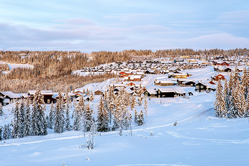 Ein weitläufiges, verschneites Hüttendorf schmiegt sich an die Hänge von Hafjell, umgeben von dicht verschneiten Nadelbäumen. Das warme Licht der tiefstehenden Wintersonne taucht die verschneite Landschaft und die Holzhäuser in eine sanfte, idyllische Atmosphäre. Skigebiete Norwegen Hafjell