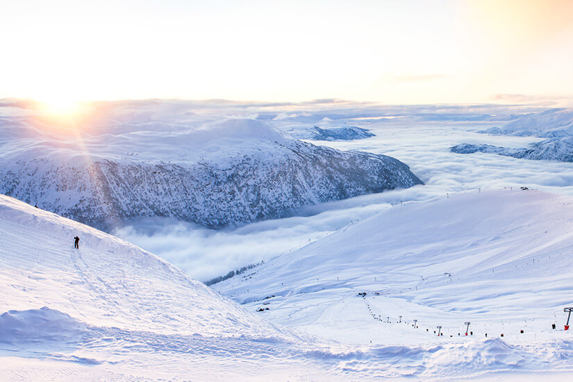 Ein einzelner Skifahrer fährt auf einem steilen, tief verschneiten Hang im Skigebiet Myrkdalen dem Sonnenuntergang entgegen. Das Tal liegt unter einer dichten, weißen Wolkendecke, aus der schneebedeckte Bergrücken hervorragend emporragen. Skigebiete Norwegen Myrkdalen