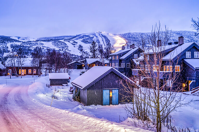 Verschneite dunkle Holzhäuser und Gebäude stehen im winterlichen Ort Geilo in der Dämmerung unter einem blauen Himmel. Im Hintergrund zieht sich eine beleuchtete Skipiste den Hang hinauf. Skigebiete Norwegen Geilo