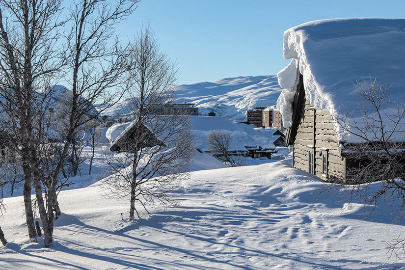 Traditionelle Holzhütten mit meterhohen, schneebedeckten Dächern stehen im Skigebiet Hovden inmitten einer tief verschneiten Landschaft. Im Hintergrund zeichnen sich sanfte, schneebedeckte Bergrücken unter einem strahlend blauen Himmel ab. Skigebiete Norwegen Hovden
