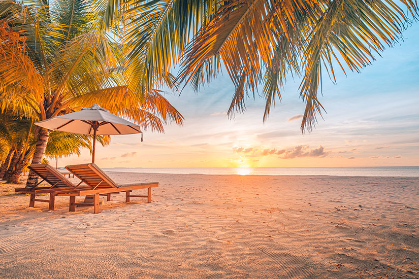 Zwei hölzerne Liegen mit einem Sonnenschirm stehen im feinen Sandstrand unter den Blättern von Palmen, während im Hintergrund die Sonne im ruhigen Meer untergeht. Pauschalreisen unter 500 Euro Entspannen