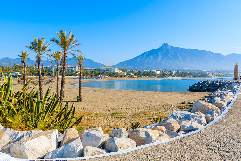 Ein heller Sandstrand mit Palmen und einer steinernen Mole erstreckt sich an der Costa del Sol, während im Hintergrund ein markanter Berg unter blauem Himmel emporragt. Pauschalreisen unter 500 Euro Costa del Sol