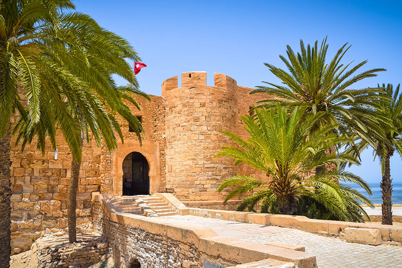 Eine historische Festung aus sandsteinfarbenem Mauerwerk auf Djerba wird von Palmen umrahmt, während ein steinerner Weg zum Eingang mit Torbogen führt. Pauschalreisen unter 500 Euro Djerba