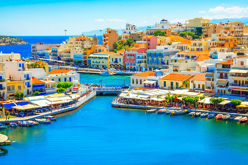 Bunte Häuser mit Terrakottadächern drängeln sich um das tiefblaue Wasser eines idyllischen Hafens in Agios Nikolaos auf Kreta. Kleine Boote liegen am Ufer vertäut, während Promenade und Restaurants zum Verweilen einladen. Pauschalreisen unter 500 Euro Kreta