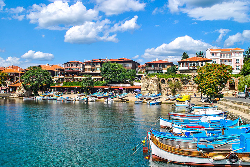 Zahlreiche kleine, blau-weiße Fischerboote liegen im ruhigen, türkisblauen Wasser eines Hafens am Sonnenstrand in Nessebar. Dahinter erstrecken sich historische Häuser mit roten Ziegeldächern unter einem blau-weißen Wolkenhimmel. Pauschalreisen unter 500 Euro Sonnenstrand