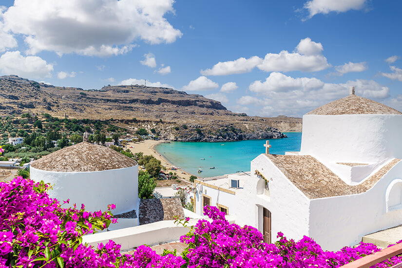 Strahlend weiße, traditionelle Kapellen mit Kuppeldächern stehen im Vordergrund, umrahmt von leuchtend pinken Bougainvillea-Blüten auf Rhodos. Im Hintergrund erstreckt sich eine geschützte Sandbucht mit türkisblauem Meer und felsigen Hügeln unter einem leicht bewölkten Himmel. Pauschalreisen unter 500 Euro Rhodos