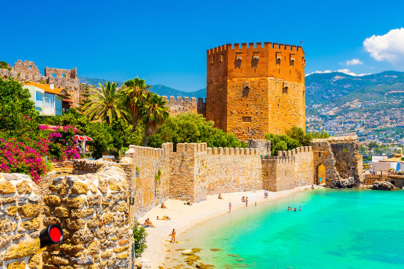 Ein wuchtiger, achteckiger Backsteinturm und eine historische Festungsmauer ragen direkt an einem feinen Sandstrand an der Türkischen Riviera empor. Das kristallklare, türkisfarbene Meer umspült den Strand, während im Hintergrund grüne Berge unter blauem Himmel aufragen. Pauschalreisen unter 500 Euro Tuerkische Riviera