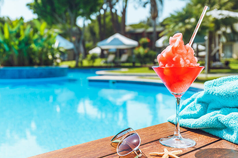 Ein rosafarbener gefrorener Cocktail in einem Cocktailglas steht zusammen mit einer Sonnenbrille, einem hellblauen Handtuch und einem Seestern auf dem Holzrand eines Swimmingpools. Im Hintergrund sind das klare blaue Poolwasser und Sonnenschirme einer Hotelanlage zu sehen. Pauschalreisen unter 500 Euro Verpflegung